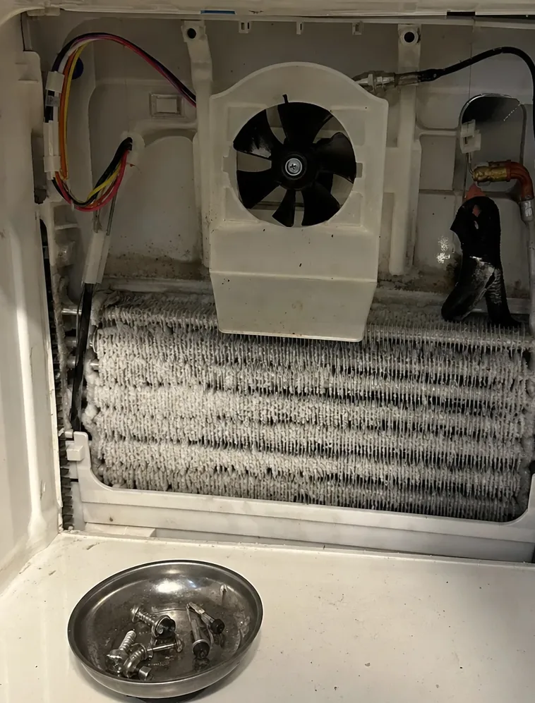 A close, straight-on view of an iced evaporator behind a removed service panel, wiring looped over the liner above.