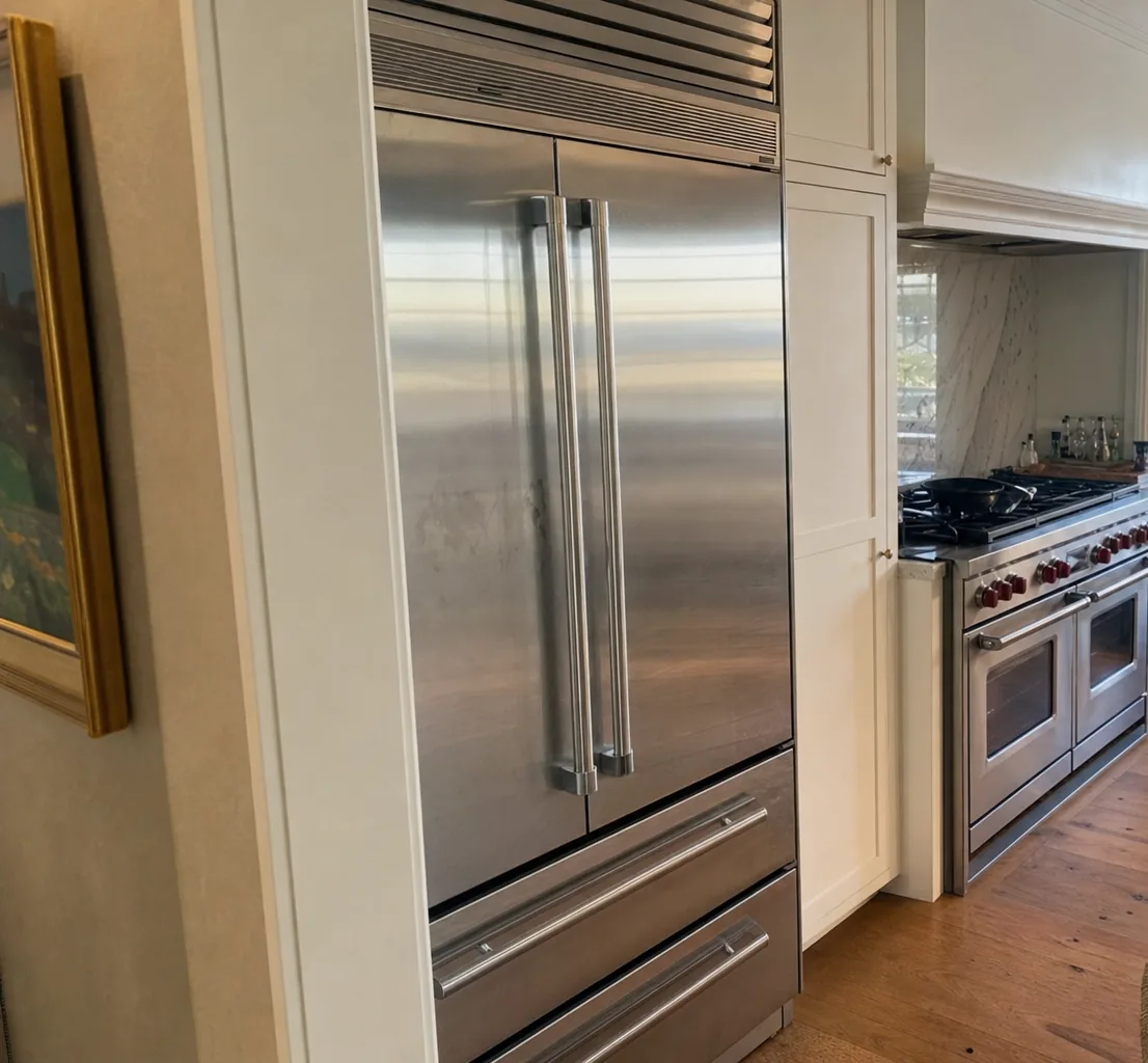 The stainless door and drawer fronts of a built-in refrigerator filling the left of a kitchen, a pro range and a bright window beyond it.