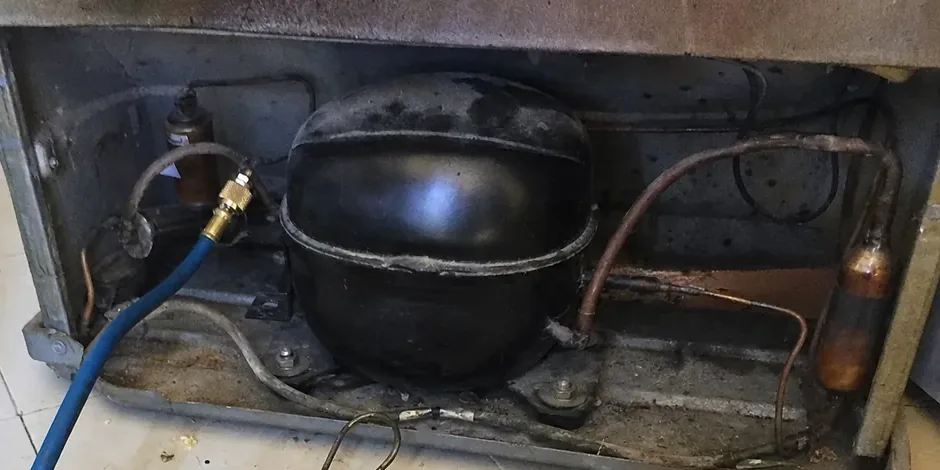 A compressor bolted into the machine compartment of a built-in, copper lines running across to a filter drier at the right and a charging hose entering from the left.